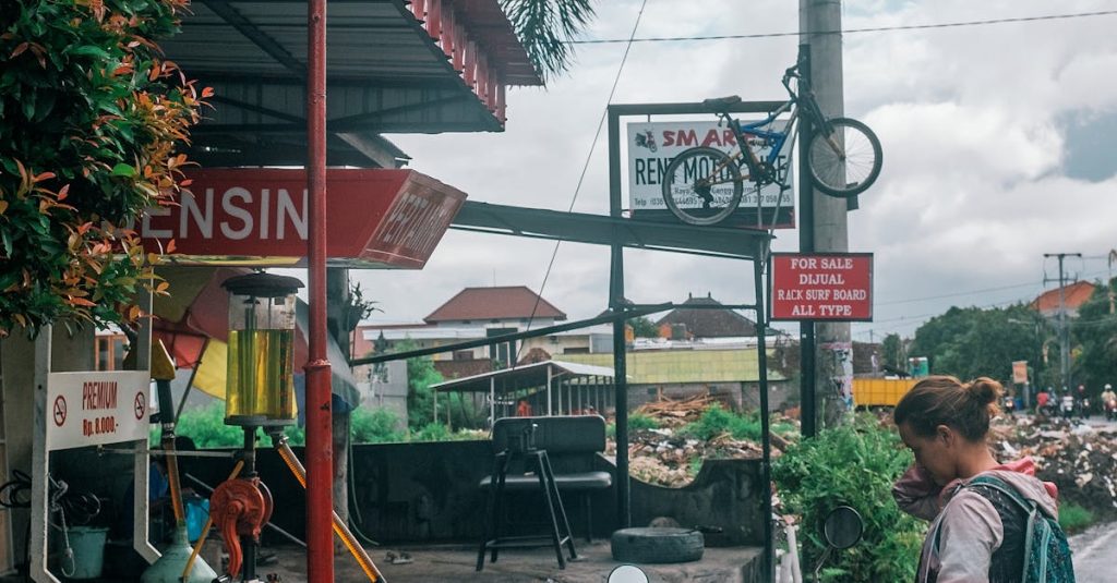 A woman standing by a motorbike at a roadside gas station in a tropical town.