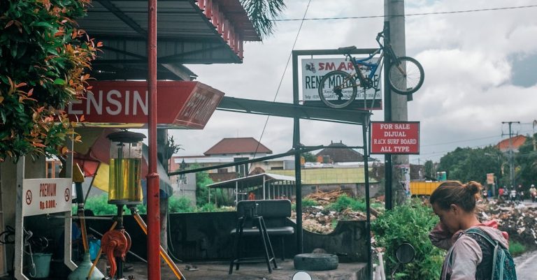 A woman standing by a motorbike at a roadside gas station in a tropical town.