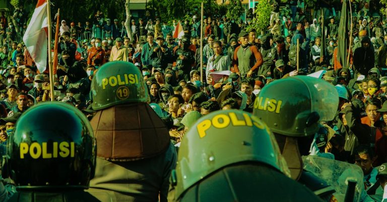 A dynamic scene of a large crowd protesting with police presence wearing helmets marked 'Polisi'.